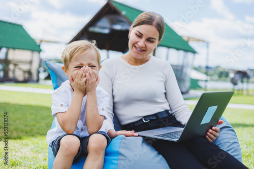 A young woman sits on a soft chair in the park and works on a laptop while her son plays next to her. Working outdoors on maternity leave.