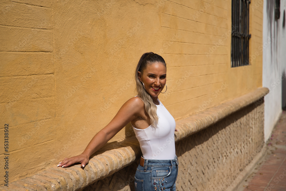 Young and beautiful woman, with ponytail, wearing a white top and jeans, smiling happily, leaning against a wall, relaxed, receiving the rays of the sun. Concept beauty, sun, relax, peace, happiness.