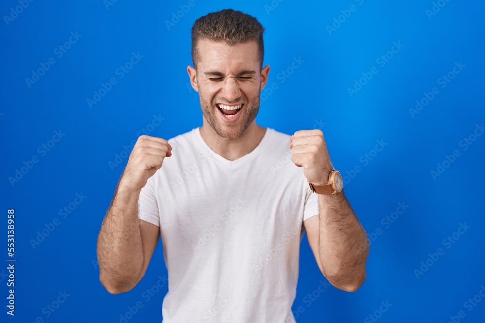 Young caucasian man standing over blue background excited for success with arms raised and eyes closed celebrating victory smiling. winner concept. Young caucasian man standing over blue background excited for success with arms raised and eyes closed celebrating victory smiling. winner concept.