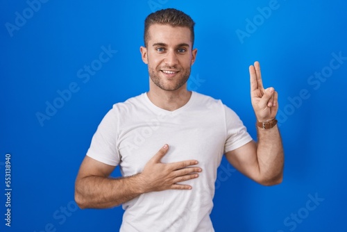 Young caucasian man standing over blue background smiling swearing with hand on chest and fingers up, making a loyalty promise oath