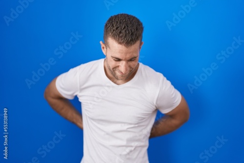 Young caucasian man standing over blue background suffering of backache, touching back with hand, muscular pain