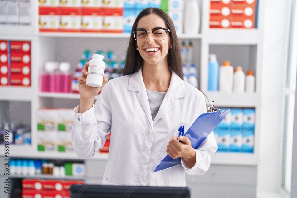 Young brunette woman working at pharmacy drugstore holding pills winking looking at the camera with sexy expression, cheerful and happy face.