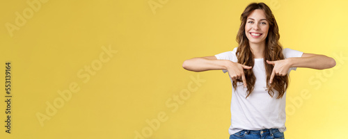 Lively helpful good-looking caucasian girl curly long hair showing you excellent place find hairdresser pointing down index fingers smiling toothy happy delighted grin stand yellow background