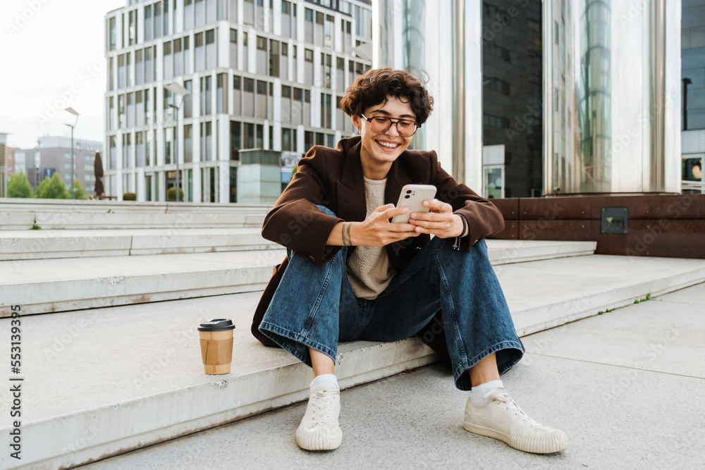 European woman using cellphone and smiling while sitting on stairs