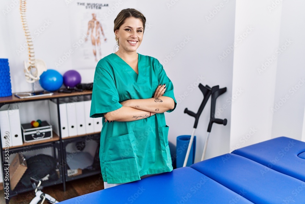 Young hispanic woman wearing physiotherapist uniform standing at clinic happy face smiling with crossed arms looking at the camera. positive person. Young hispanic woman wearing physiotherapist uniform standing at clinic happy face smiling with crossed arms looking at the camera. positive person.
