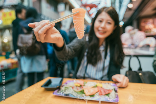 selective focus of a piece of fresh pink Japanese food raw fish sashimi held on chopsticks by an asian female diner at table in kuromon ichiba market in Osaka japan photo