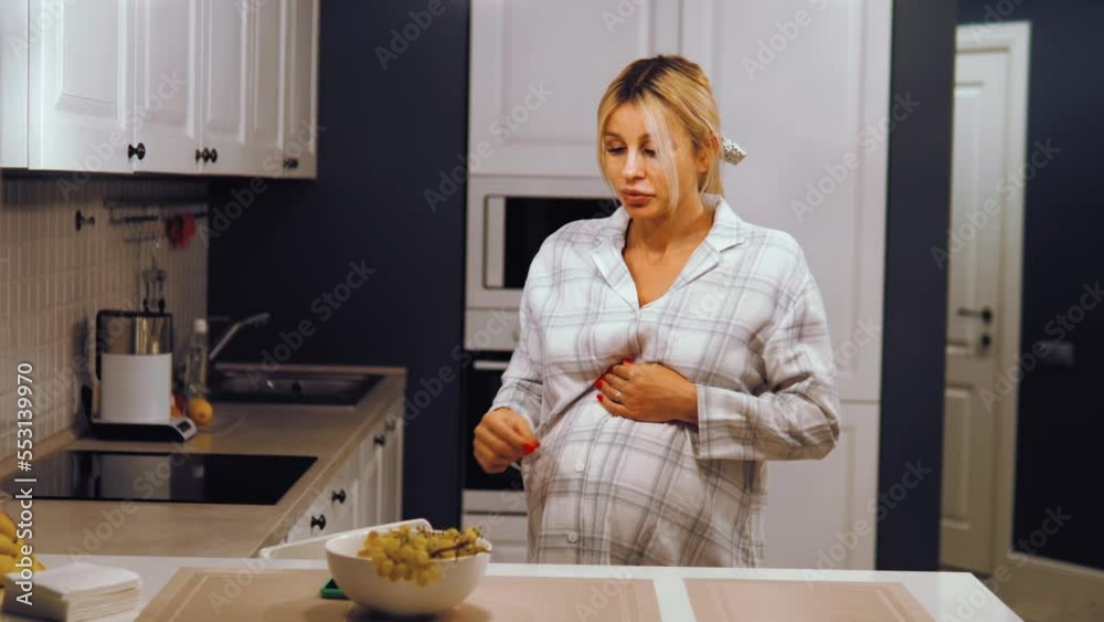 Pregnant female standing in the kitchen eating fruits grapes in the plate, hungry expectant mother with belly having snack Pregnant female standing in the kitchen eating fruits grapes in the plate, hungry expectant mother with belly having snack