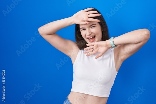 Young caucasian woman standing over blue background smiling cheerful playing peek a boo with hands showing face. surprised and exited