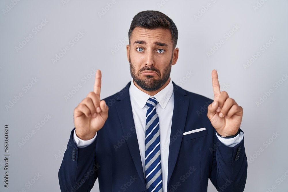 Handsome hispanic man wearing suit and tie pointing up looking sad and upset, indicating direction with fingers, unhappy and depressed. Handsome hispanic man wearing suit and tie pointing up looking sad and upset, indicating direction with fingers, unhappy and depressed.