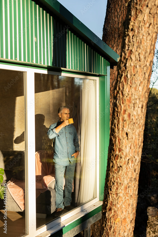 Mature man standing at the window of a house with cup of coffee looking out Mature man standing at the window of a house with cup of coffee looking out