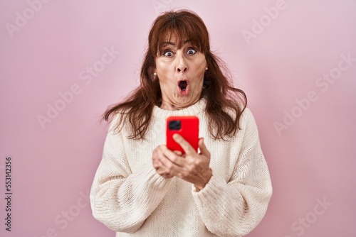 Middle age hispanic woman using smartphone typing message afraid and shocked with surprise and amazed expression, fear and excited face.
