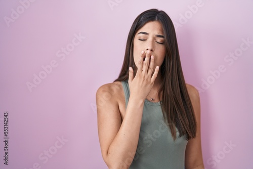 Hispanic woman standing over pink background bored yawning tired covering mouth with hand. restless and sleepiness.