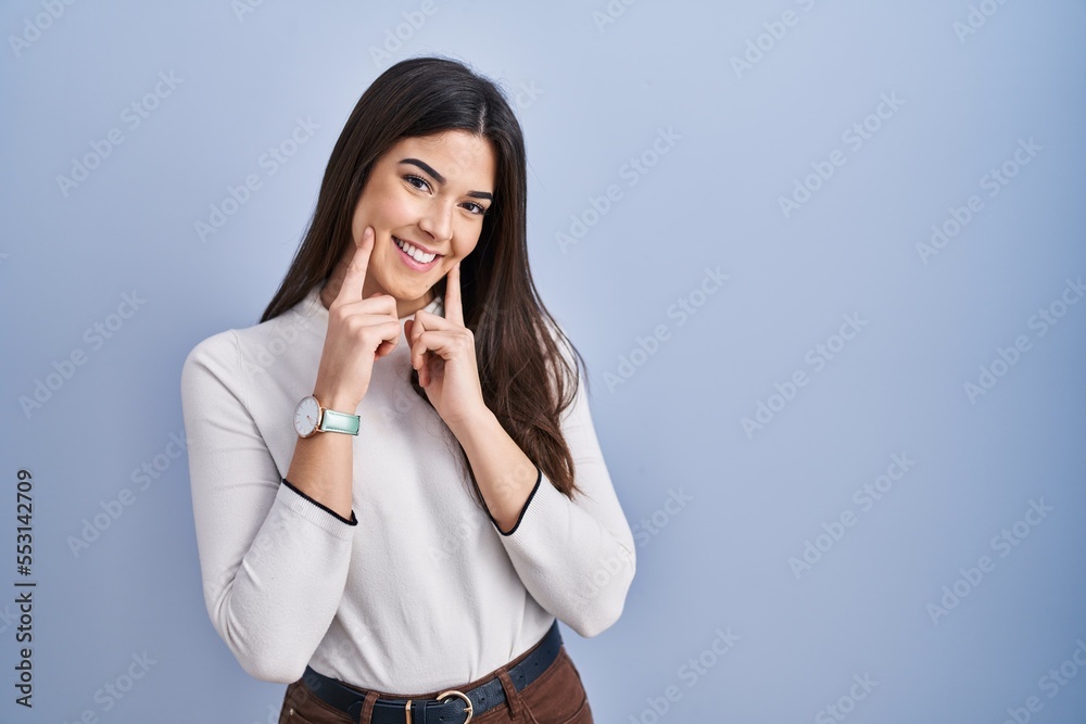 Young brunette woman standing over blue background smiling with open mouth, fingers pointing and forcing cheerful smile Young brunette woman standing over blue background smiling with open mouth, fingers pointing and forcing cheerful smile