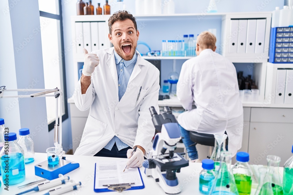 Hispanic man and woman working at scientist laboratory pointing thumb up to the side smiling happy with open mouth Hispanic man and woman working at scientist laboratory pointing thumb up to the side smiling happy with open mouth