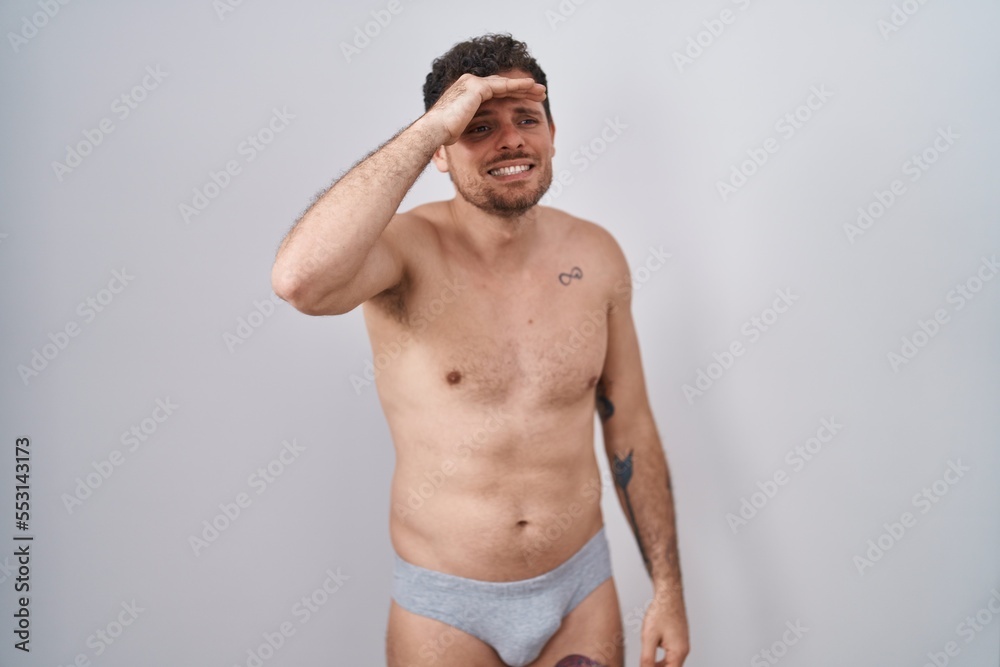 Fototapeta premium Young hispanic man standing shirtless wearing underware very happy and smiling looking far away with hand over head. searching concept.