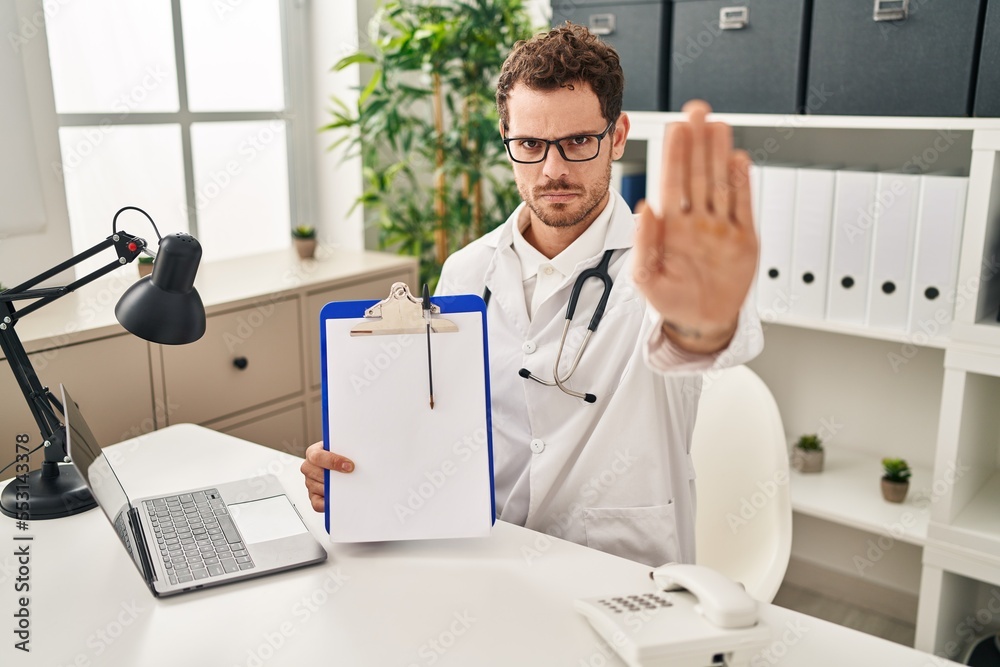 Young hispanic man wearing doctor stethoscope holding clipboard with open hand doing stop sign with serious and confident expression, defense gesture Young hispanic man wearing doctor stethoscope holding clipboard with open hand doing stop sign with serious and confident expression, defense gesture