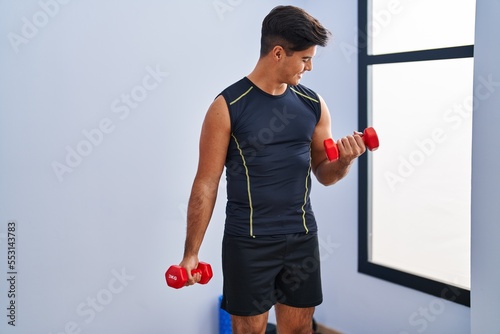 Young hispanic man smiling confident using dumbbells training at sport center