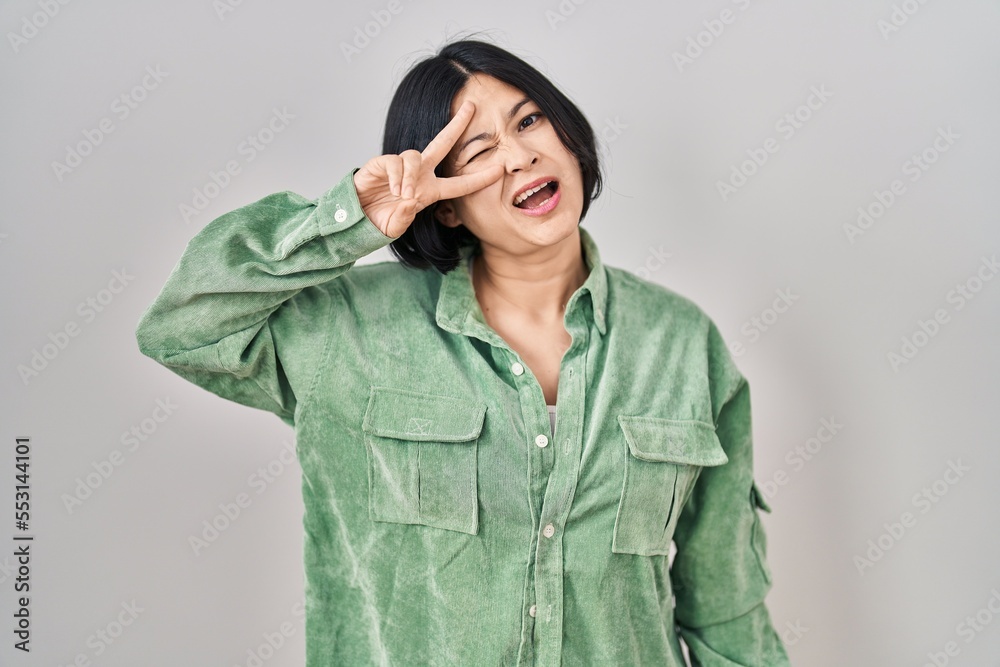 Young asian woman standing over white background doing peace symbol with fingers over face, smiling cheerful showing victory Young asian woman standing over white background doing peace symbol with fingers over face, smiling cheerful showing victory