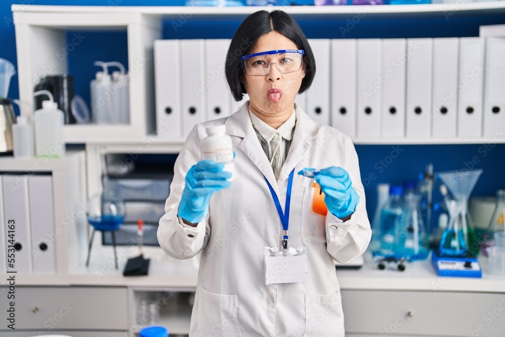 Young asian woman working at scientist laboratory sticking tongue out happy with funny expression.