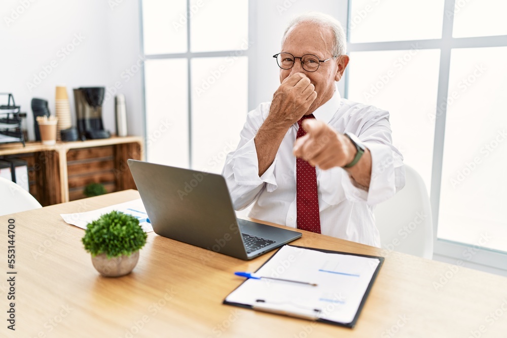 Senior man working at the office using computer laptop laughing at you, pointing finger to the camera with hand over mouth, shame expression