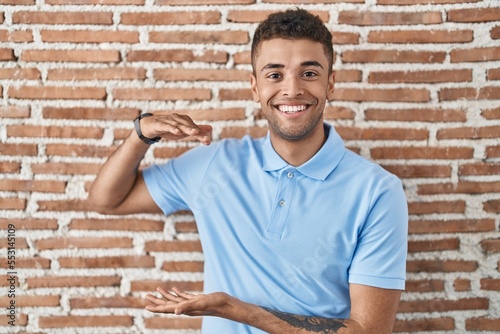 Brazilian young man standing over brick wall gesturing with hands showing big and large size sign, measure symbol. smiling looking at the camera. measuring concept.