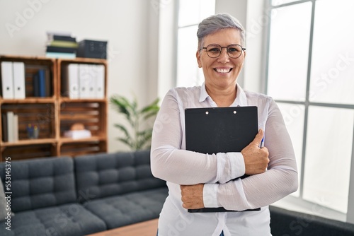 Middle age woman psychologist holding clipboard standing at pyschology center photo