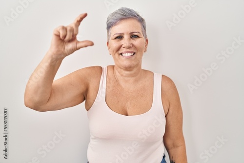 Middle age caucasian woman standing over white background smiling and confident gesturing with hand doing small size sign with fingers looking and the camera. measure concept.