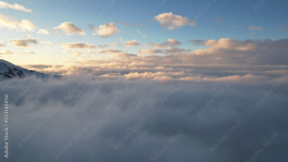 Flying among the ocean of clouds in the mountains. The fog is like big ocean waves crashing against high mountains covered with snow. Spruce trees grow in places. Sunset. Orange rays of the sun