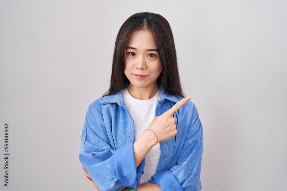 Young chinese woman standing over white background pointing with hand finger to the side showing advertisement, serious and calm face