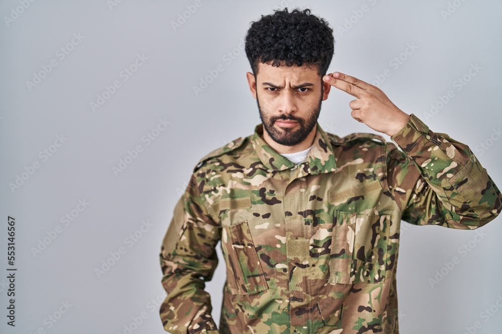 Arab man wearing camouflage army uniform shooting and killing oneself pointing hand and fingers to head like gun, suicide gesture.