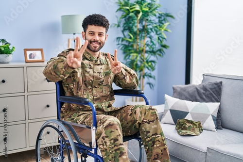 Arab man wearing camouflage army uniform sitting on wheelchair smiling looking to the camera showing fingers doing victory sign. number two.