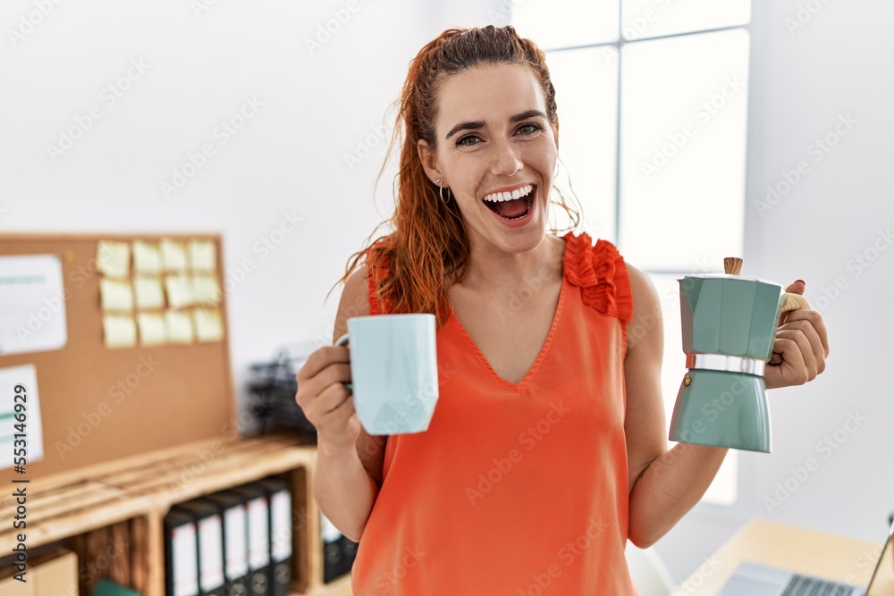 Young redhead woman holding french coffee maker at the office smiling and laughing hard out loud because funny crazy joke. Young redhead woman holding french coffee maker at the office smiling and laughing hard out loud because funny crazy joke.