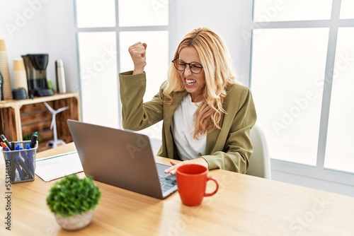 Beautiful blonde woman working at the office with laptop angry and mad raising fist frustrated and furious while shouting with anger. rage and aggressive concept.