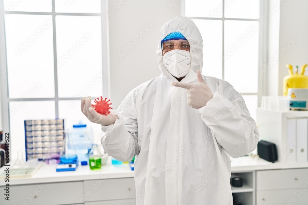 Hispanic young man working at scientist laboratory holding virus toy pointing finger to one self smiling happy and proud Hispanic young man working at scientist laboratory holding virus toy pointing finger to one self smiling happy and proud