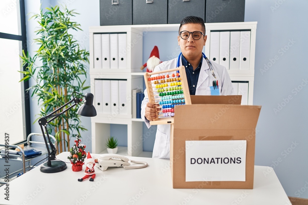 Hispanic young doctor man donating toys at the clinic relaxed with serious expression on face. simple and natural looking at the camera. Hispanic young doctor man donating toys at the clinic relaxed with serious expression on face. simple and natural looking at the camera.