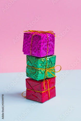 Colorful gifts wrapped in shiny packaging for Christmas or New Year are stacked on a pink and blue background.