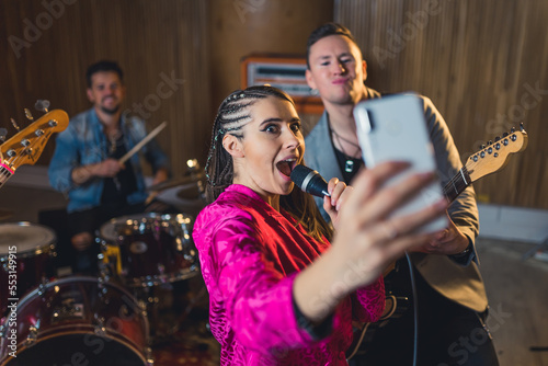 Spontaneous funky woman singer takes a selfie with white phone with her band members during an indoor concert. High quality photo