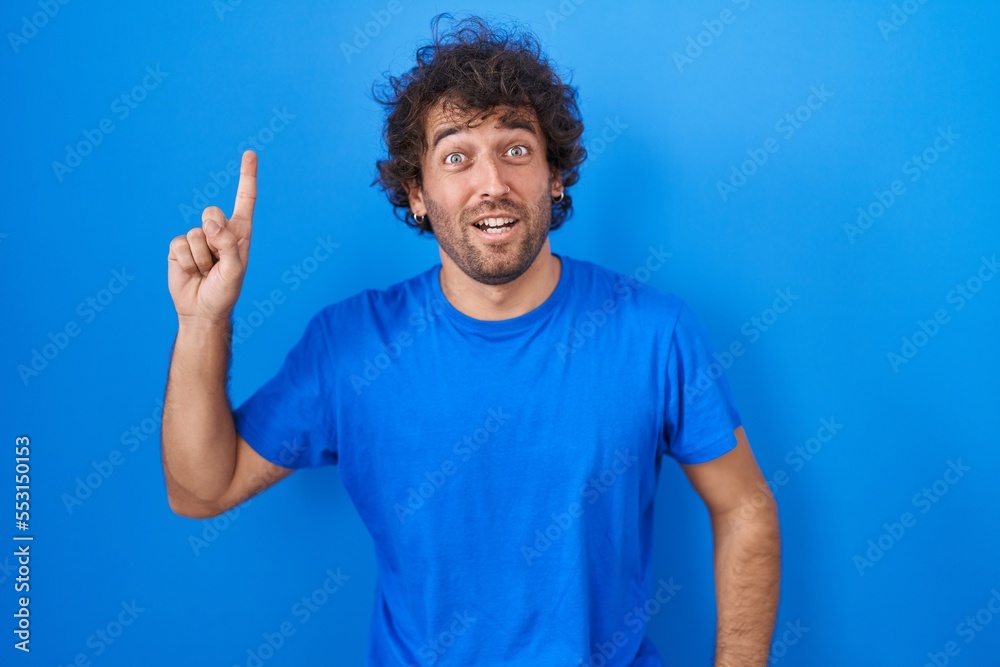Naklejka premium Hispanic young man standing over blue background pointing finger up with successful idea. exited and happy. number one.