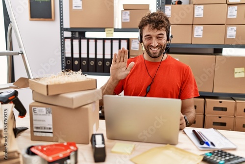 Young hispanic call center agent man working at warehouse waiving saying hello happy and smiling, friendly welcome gesture