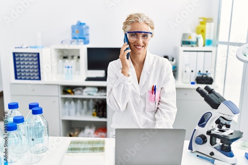 Middle age blonde woman wearing scientist uniform talking on the smartphone working at laboratory