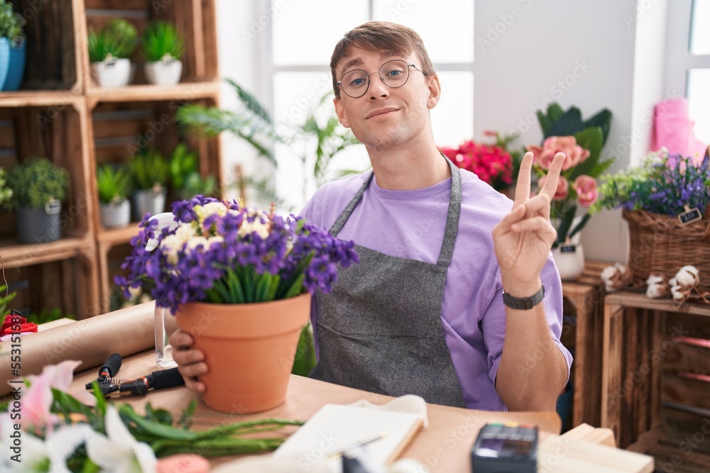 Caucasian blond man working at florist shop smiling with happy face winking at the camera doing victory sign with fingers. number two.