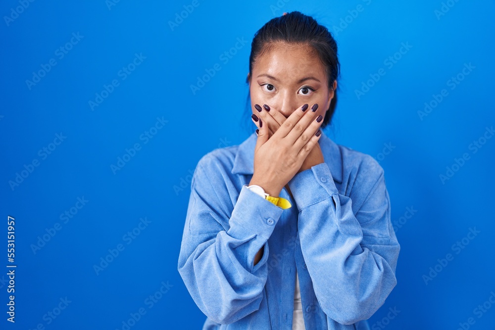 Asian young woman standing over blue background shocked covering mouth with hands for mistake. secret concept. Asian young woman standing over blue background shocked covering mouth with hands for mistake. secret concept.