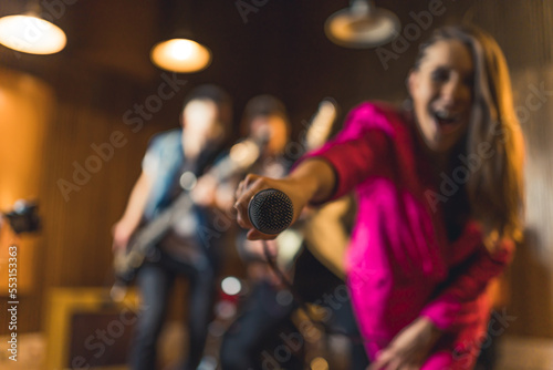 Camera focus on microphone. Blurred excited female singer in pink suit and guitarists in the background. Band and performance. High quality photo