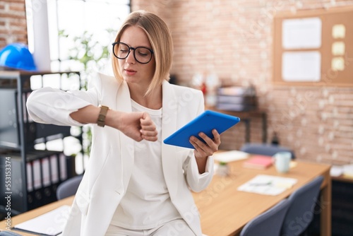 Young caucasian woman working at the office wearing glasses checking the time on wrist watch, relaxed and confident