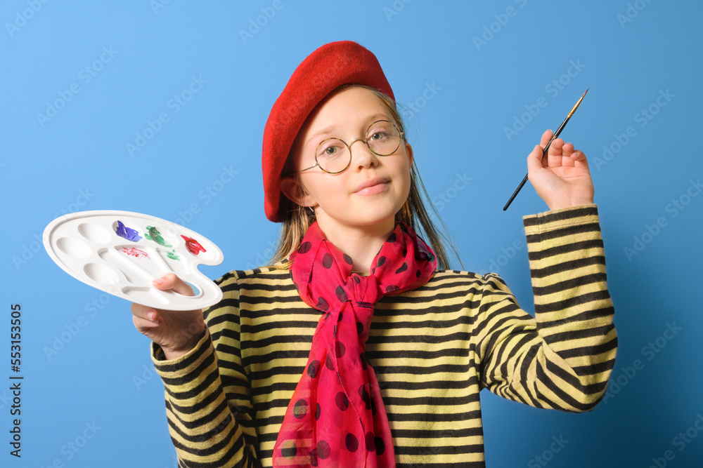 Fototapeta premium Young artist. A girl with a palette of colors in a bright red beret and with a brush on a blue background. close-up portrait