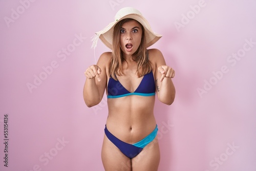 Young hispanic woman wearing bikini over pink background pointing down with fingers showing advertisement, surprised face and open mouth