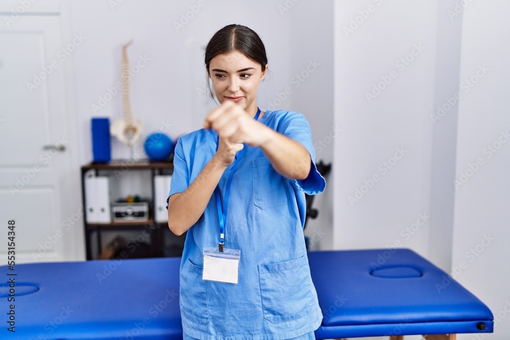 Obraz premium Young hispanic woman wearing physiotherapist uniform standing at clinic punching fist to fight, aggressive and angry attack, threat and violence