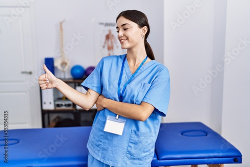 Young hispanic woman wearing physiotherapist uniform standing at clinic looking proud, smiling doing thumbs up gesture to the side