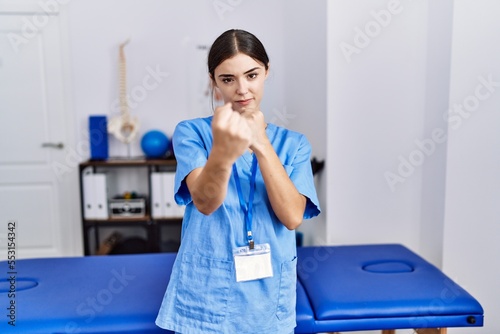 Young hispanic woman wearing physiotherapist uniform standing at clinic ready to fight with fist defense gesture  angry and upset face  afraid of problem