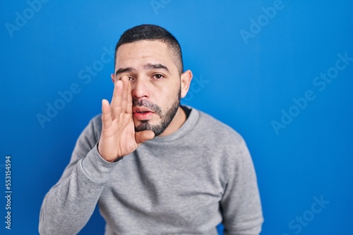 Hispanic man standing over blue background hand on mouth telling secret rumor, whispering malicious talk conversation © Krakenimages.com